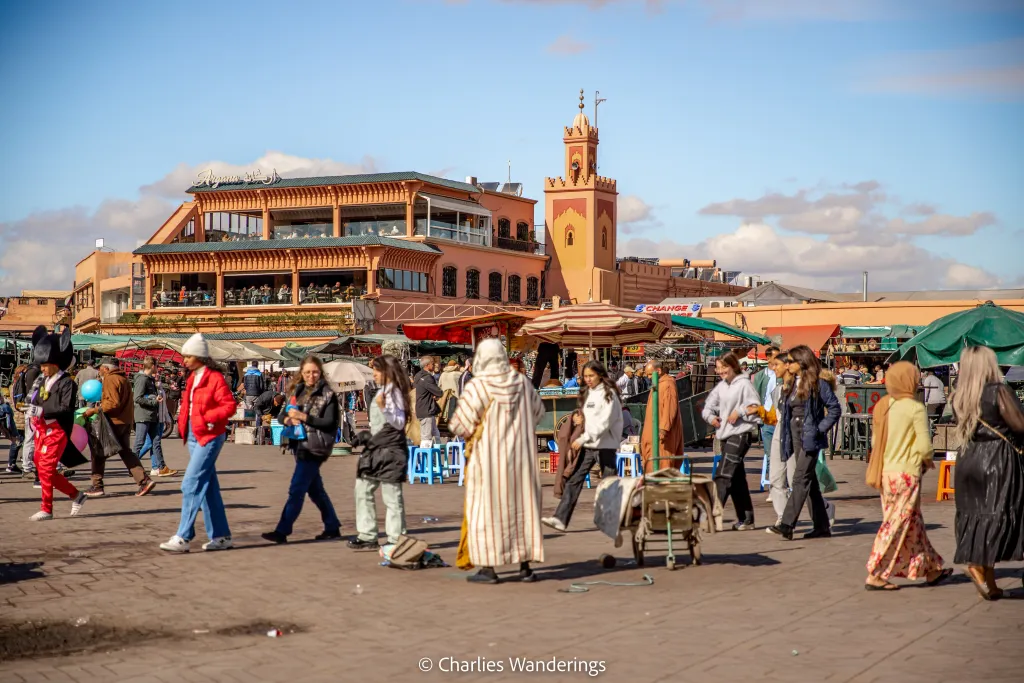 Bustling Jemaa el-Fnaa square in Marrakech, Morocco at the height of day. A vibrant scene filled with local merchants, snake charmers, acrobats, and food stalls. Intricate architecture of the surrounding buildings frames the lively marketplace, with dappled sunlight filtering through the awnings and canopies. In the foreground, a sea of colorful textiles, spices, and handicrafts tempt passersby. Mid-ground bustles with energetic crowds, while in the background, the iconic minaret of the Koutoubia Mosque rises majestically. An atmosphere of immersive cultural exchange, sights, sounds, and aromas permeate the historic heart of the Red City. jemaa el-fnaa marrakech town centre